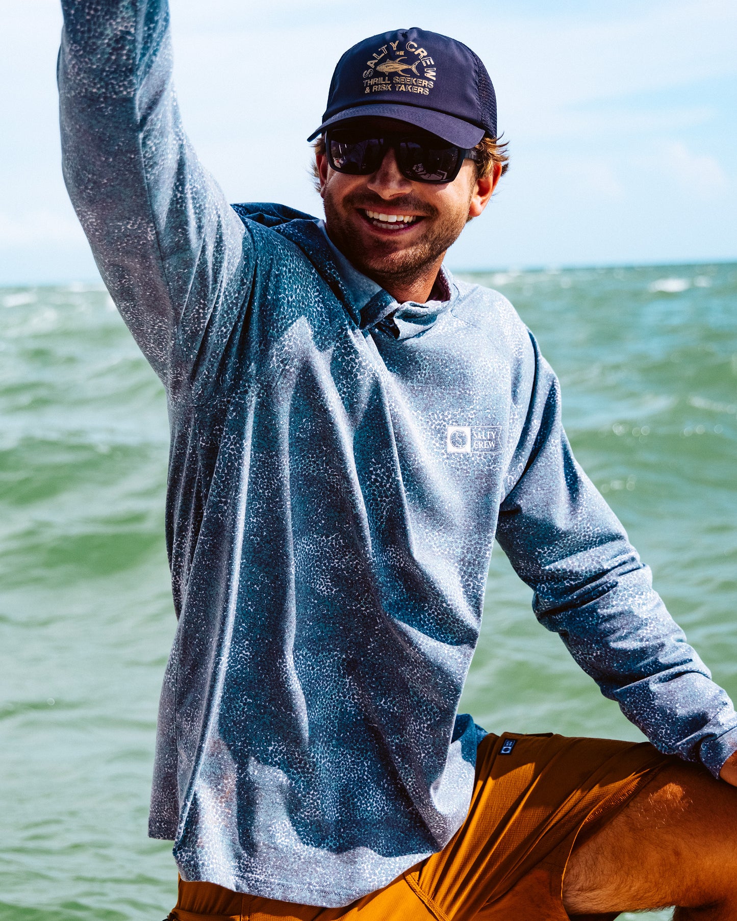 A smiling man wears a Salty Crew Mariner Lite UV Hoodie in Dark Navy, orange shorts, sunglasses, and a navy cap as he stands by the ocean, raising one arm joyfully with waves and blue sky behind him.