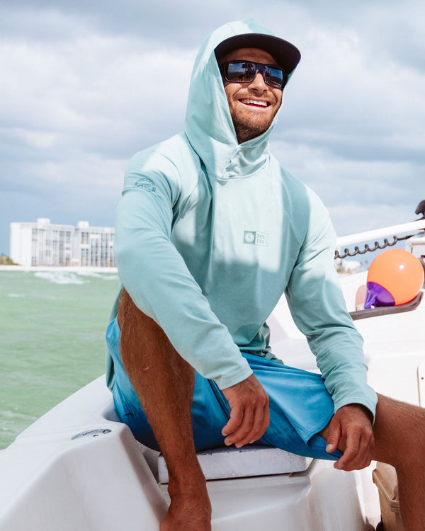 A man wearing sunglasses and a Mariner Lite UV Hoodie - Marine, along with blue shorts, smiles while sitting on a boat. In the background, water and some buildings are visible beneath a partly cloudy sky.