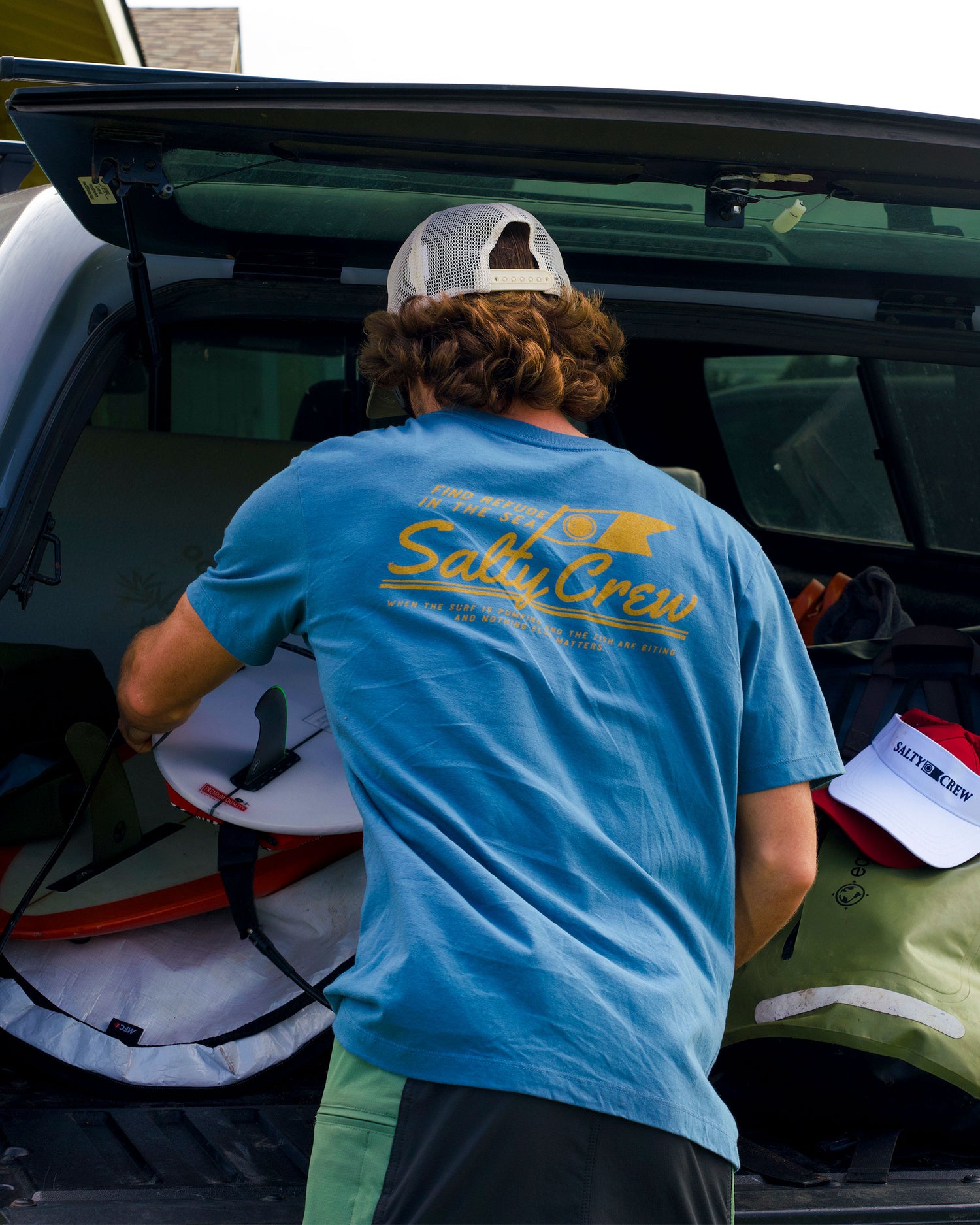 A man in a Salty Crew Original Tee - Slate and a mesh cap loads surfboards and gear into an SUV, suggesting he's getting ready for a surfing trip.