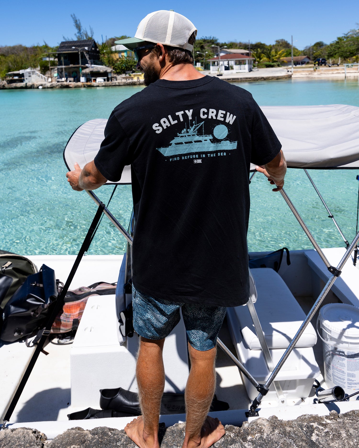 A man in a black Salty Crew Overnight Tee and patterned shorts stands on a rocky shoreline beside a docked boat in clear turquoise water, adjusting the boat's canopy under the sun.
