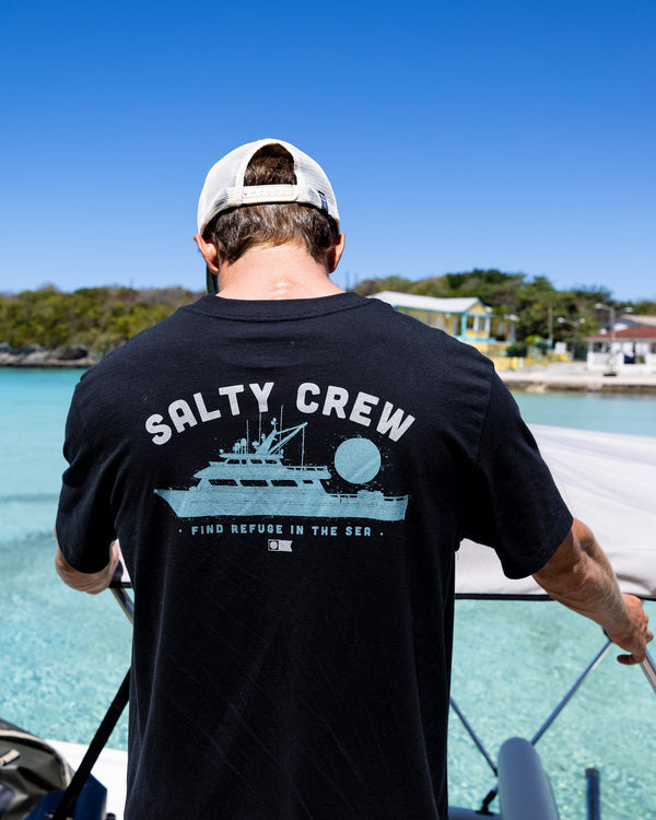 A man in a Salty Crew Overnight Tee - Black and a backwards cap stands by clear blue water, holding a boat railing, with houses and trees behind him under a bright blue sky.