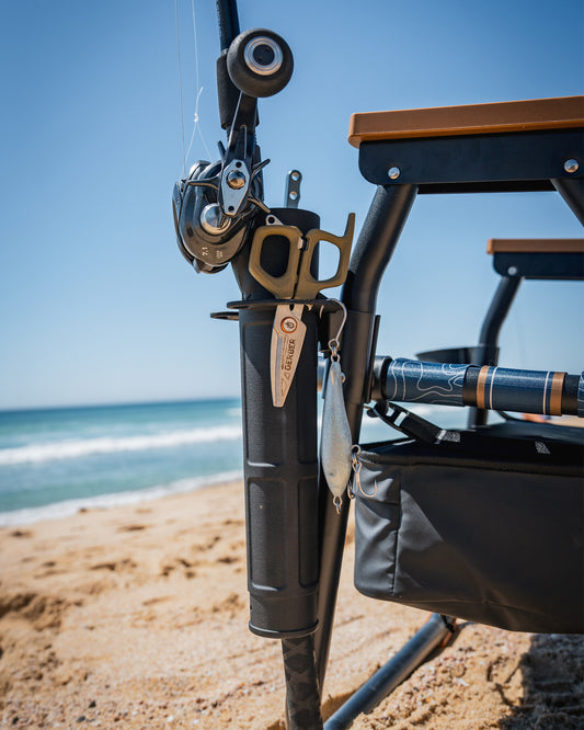 A close-up of the Parkit x Salty Crew Voyager Accessory Mount by PARKIT Co. on a sandy beach, displaying fishing gear like a rod holder, reel, pliers, and lure attached to the chair with the ocean and blue sky behind.