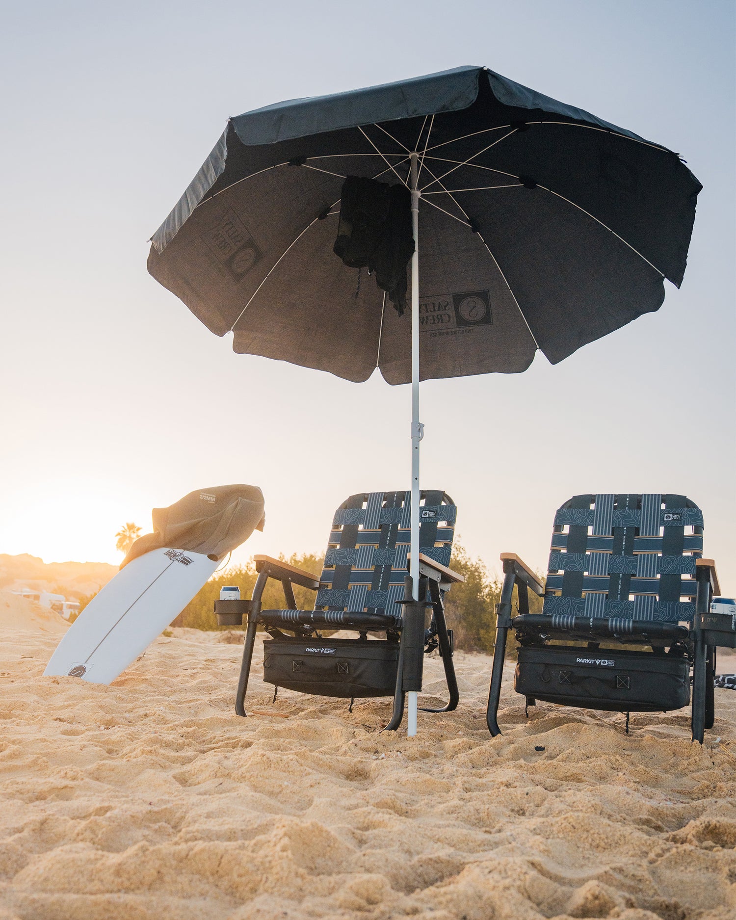 Two empty beach chairs sit under a large black umbrella at sunset, with a PARKIT Co. Parkit x Salty Crew Voyager Accessory Mount and a white surfboard propped nearby. Sparse greenery is visible in the background.