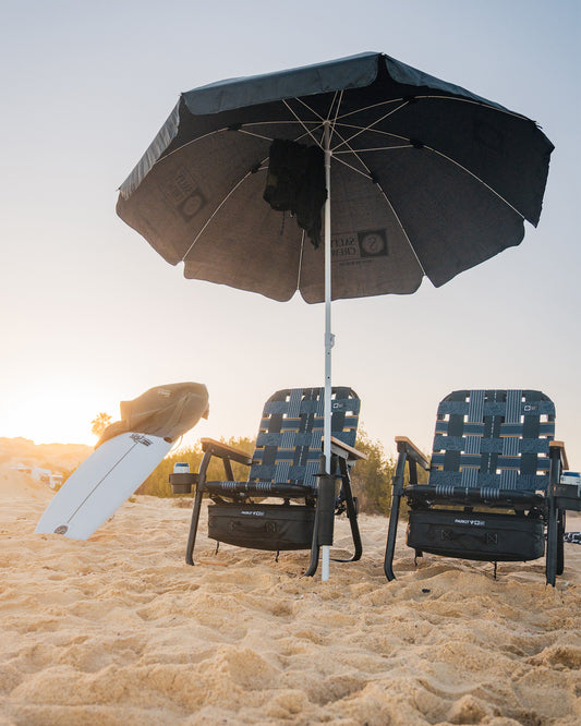Two empty beach chairs sit under a large black umbrella at sunset, with a PARKIT Co. Parkit x Salty Crew Voyager Accessory Mount and a white surfboard propped nearby. Sparse greenery is visible in the background.