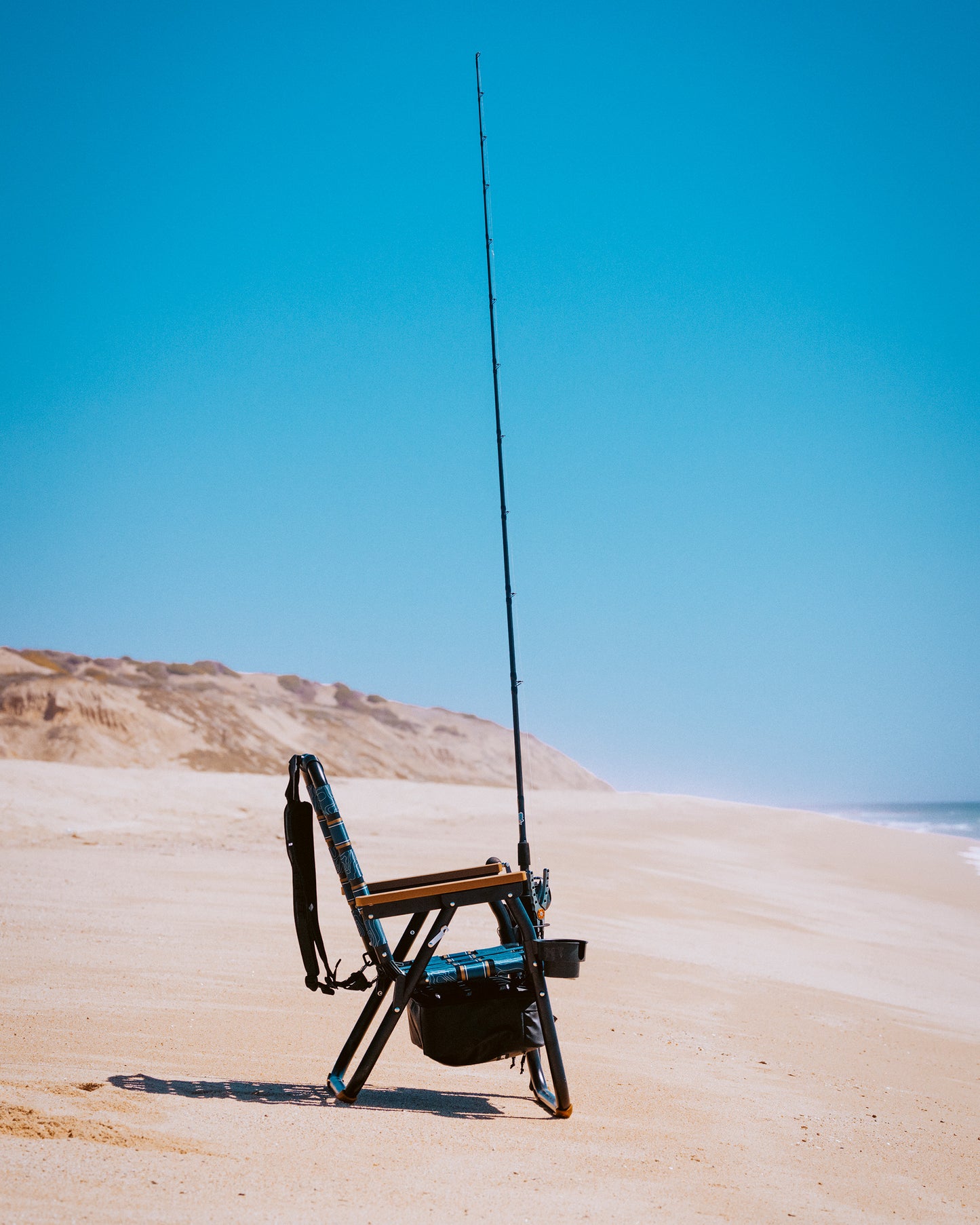 The PARKIT Co. Parkit x Salty Crew Voyager Accessory Mount, with a fishing rod propped beside it, rests on a sandy beach under clear blue skies, dunes in the distance and shoreline stretching out—your perfect fishing chair companion.