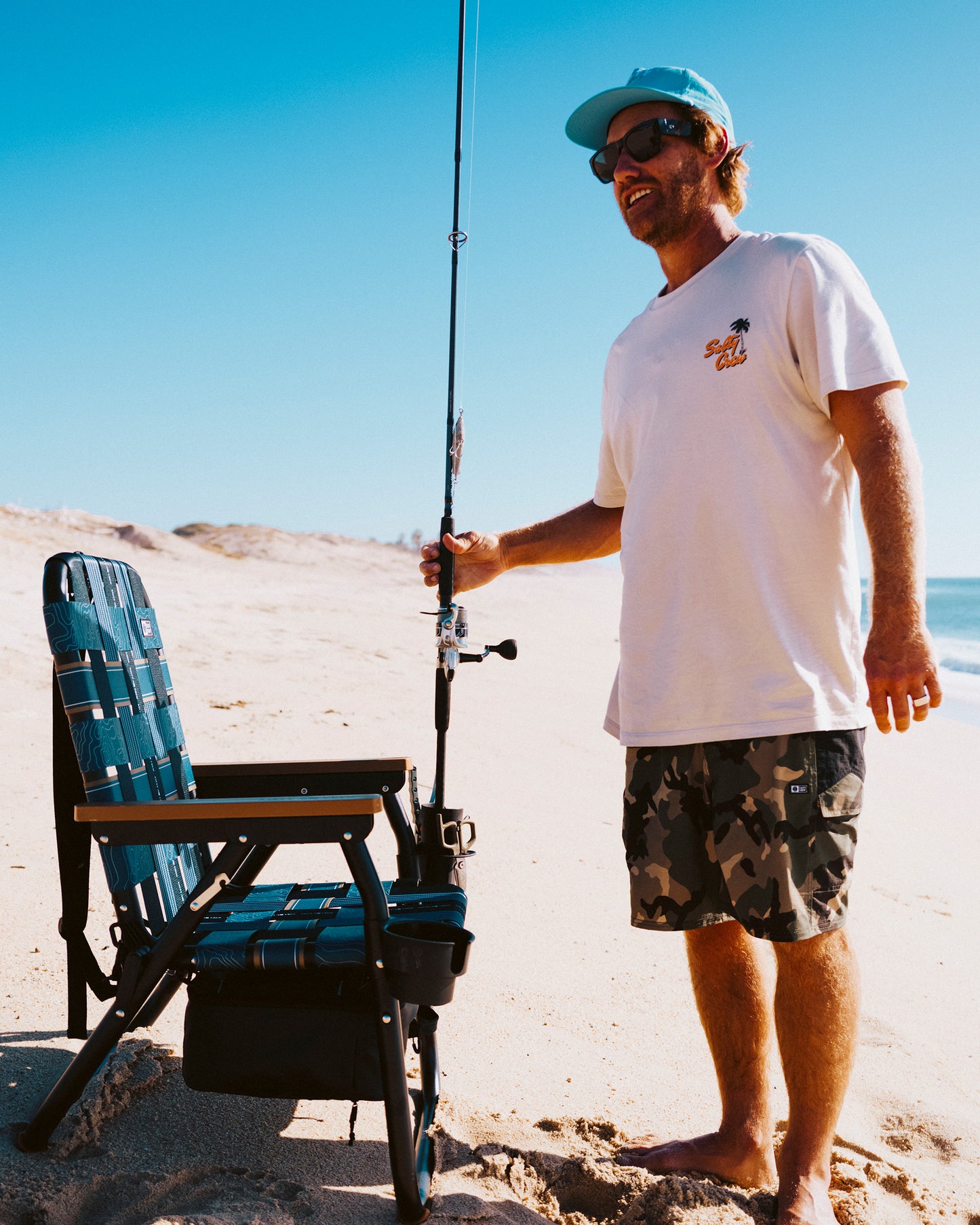 A man in sunglasses, blue cap, white t-shirt, and camo shorts stands on a sunny beach holding a fishing rod beside his folding chair fitted with the PARKIT Co. Parkit x Salty Crew Voyager Accessory Mount.