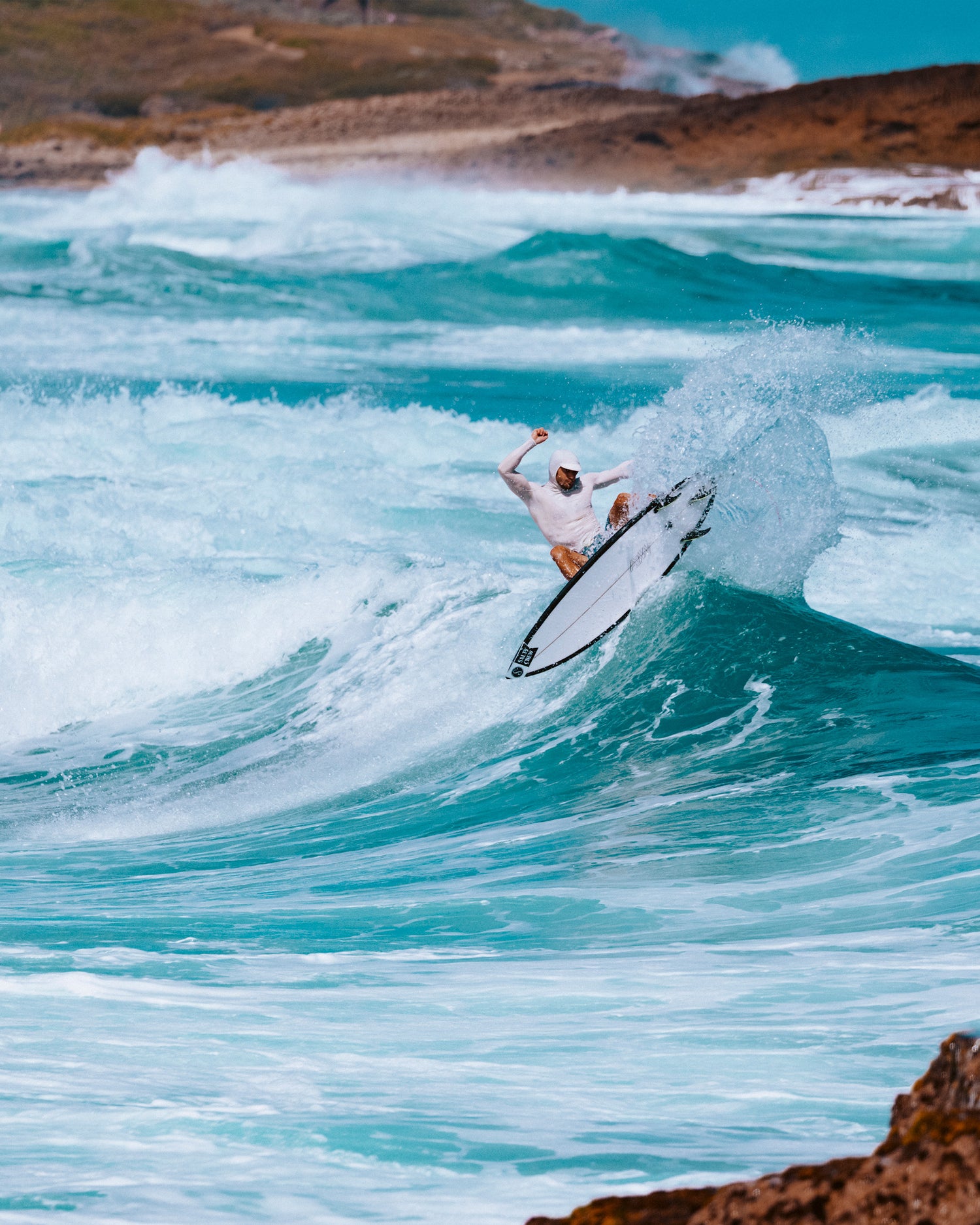 A surfer in the Salty Crew Apex Hood Surf Rashguard - Light Grey rides a turquoise wave near rocky shore, ocean in the background and white water spraying around the surfboard.