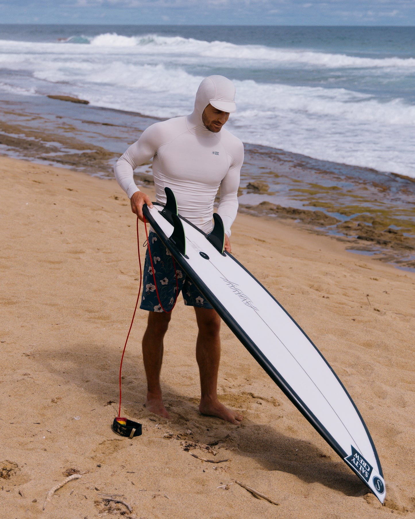 A person in a Salty Crew Apex Hood Surf Rashguard - Light Grey and patterned board shorts stands barefoot on a sandy beach by the ocean, holding a surfboard with waves and rocks in the background.