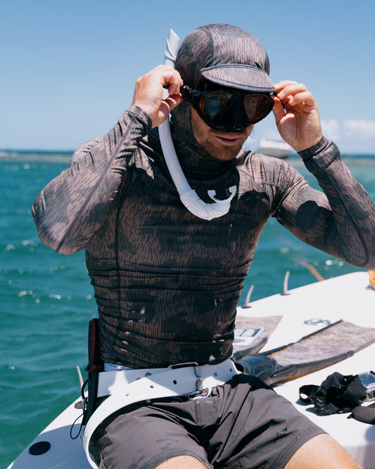 Wearing the Salty Crew Apex Hooded Rashguard - Recon Camo with UPF 50+ and a snorkel mask, a person adjusts their goggles on a boat, with the ocean and blue sky stretching out behind them.