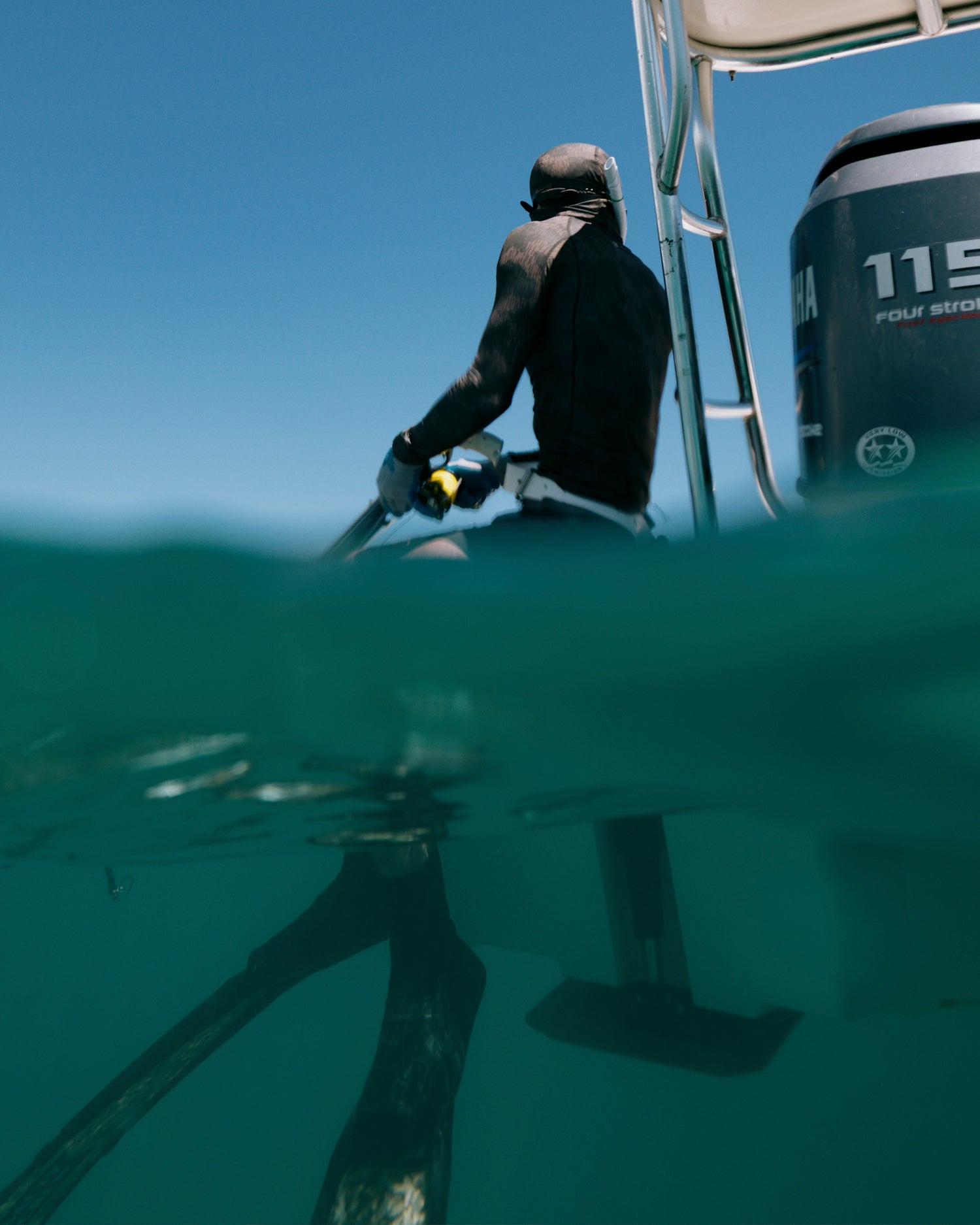 Wearing the Salty Crew Apex Hooded Rashguard - Recon Camo with UPF 50+, a snorkeler in fins climbs a boat ladder from the ocean as a Yamaha 115 outboard gleams under the clear blue sky.