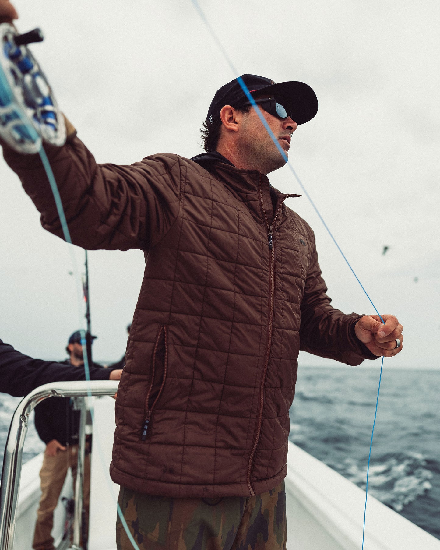 A man wearing a deep brown Salty Crew Barrier Lite Jacket and baseball cap stands on a boat, holding a fishing line.