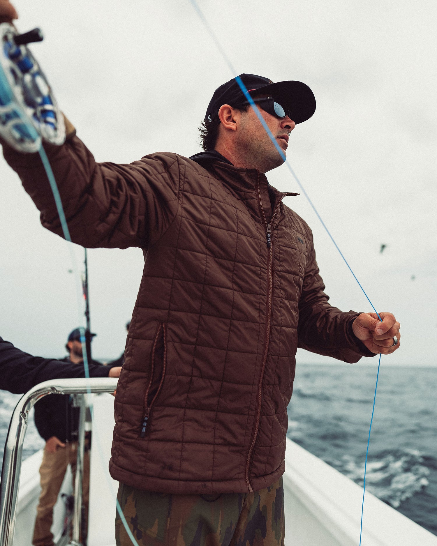 A man wearing a deep brown Salty Crew Barrier Lite Jacket and baseball cap stands on a boat, holding a fishing line.