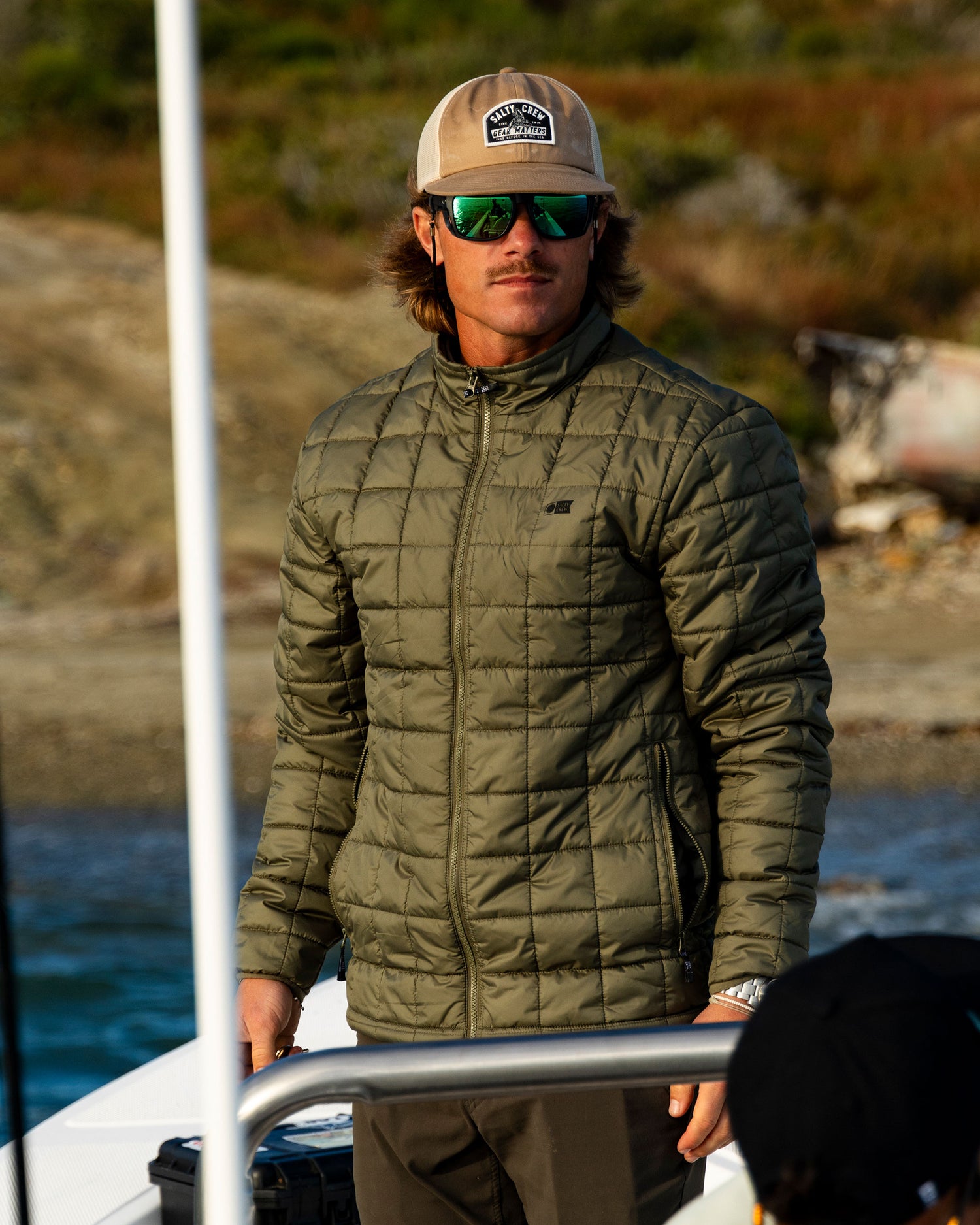 A man wearing a green Salty Crew Barrier Lite Jacket - Washed Leaf, a baseball cap, and sunglasses stands on a boat.
