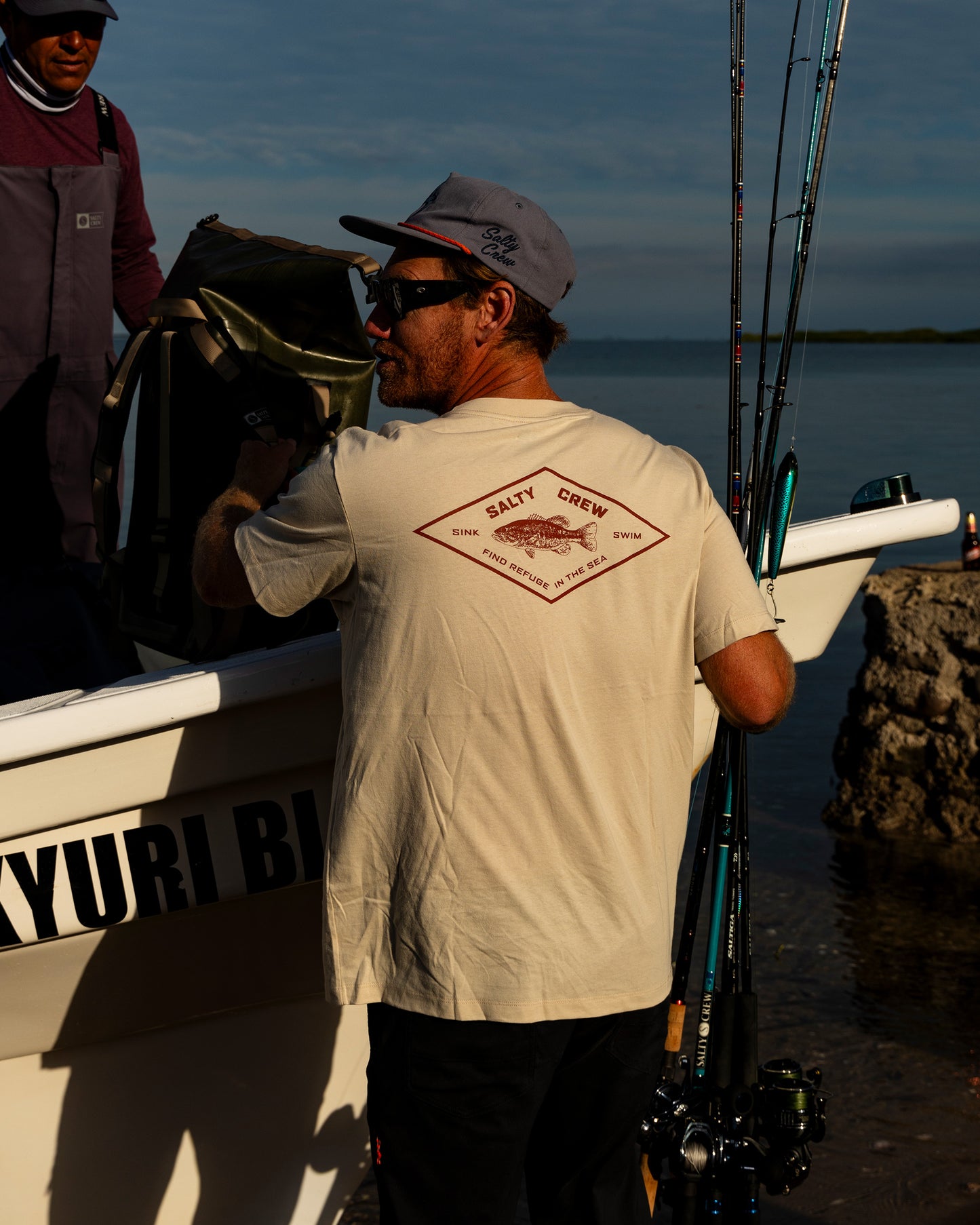 A man in sunglasses and a cap stands beside a boat holding fishing rods. He wears the Salty Crew Diamond Bass Crew Tee in Bone, a cotton t-shirt screenprinted with the brand’s logo. Another person is partly visible. The scene is set by water, with blue sky and rocks in the background.