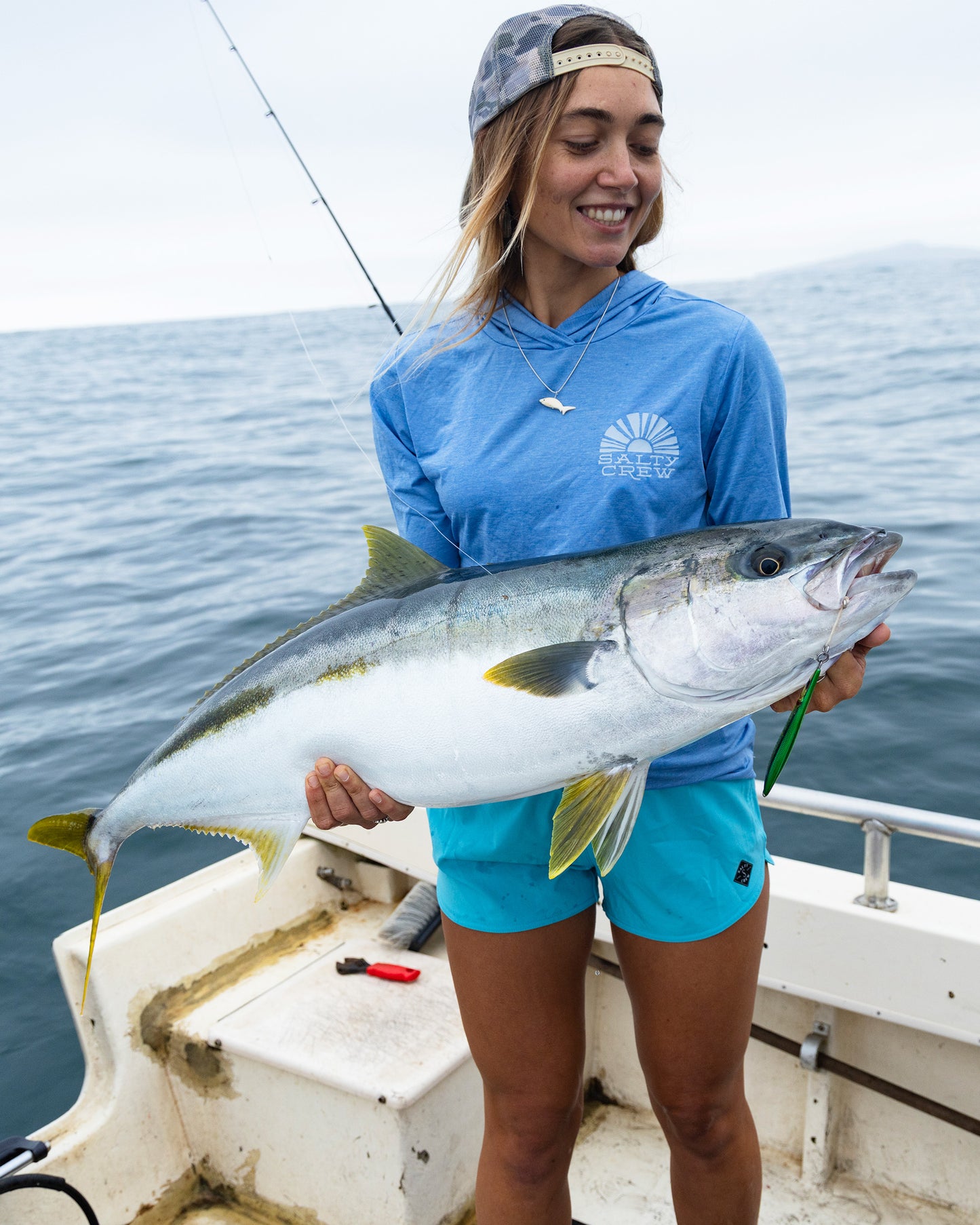 A woman wearing the Salty Crew Beacons Short in Turquoise smiles while holding a large fish on a boat, with the ocean and a fishing rod in the background.