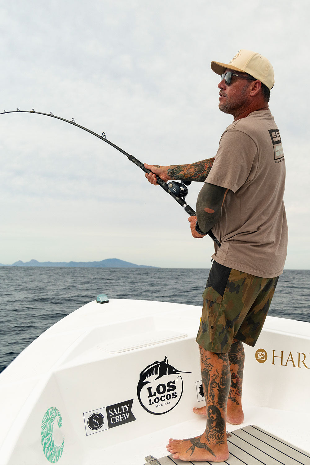 A tattooed man in sunglasses stands barefoot on a boat, holding a fishing rod over the water. He’s outfitted in Salty Crew’s Flagship 20″ Boatshort - Ranger, looking ready for anything as he patiently waits for a catch. Stickers like Los Locos decorate the boat, and mountains rise in the distance beneath a cloudy sky.