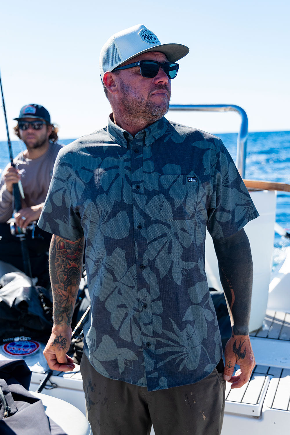 A man wearing Salty Crew’s Breezer Perforated Button Up in Black/Green, along with sunglasses and a cap, stands on a boat with the ocean in the background while another person sits behind him holding a fishing rod.
