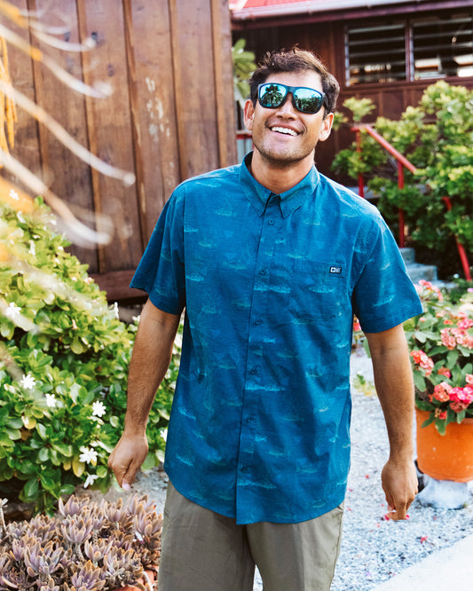 A man wearing the Salty Crew Breezer Perforated Button Up in navy stands outside, smiling amid greenery and flowers with a wooden building behind him. The lightweight, breathable shirt is perfect for warm weather.