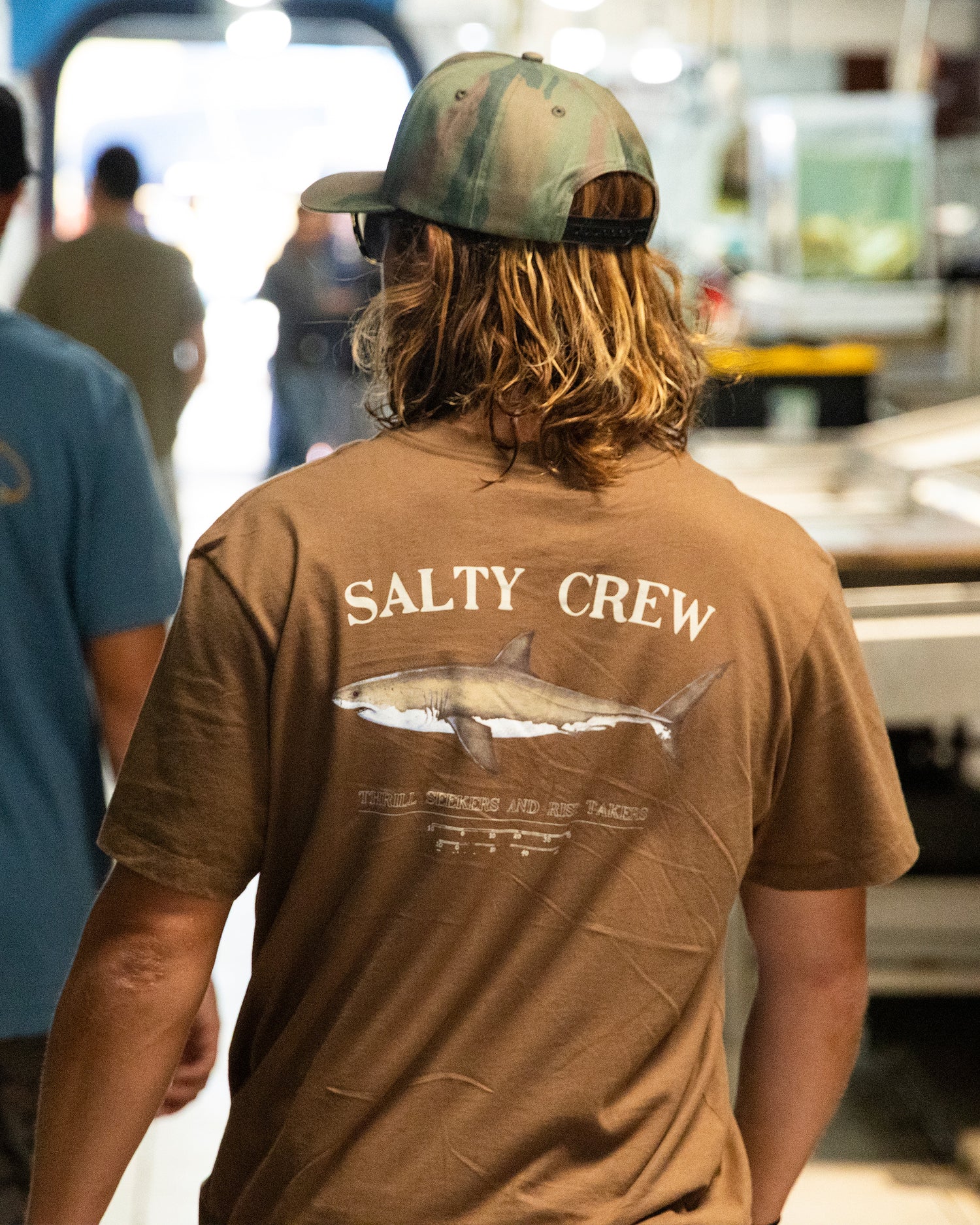 A person with long hair is seen from behind indoors, wearing the Salty Crew Bruce Crew Tee in Sepia—a brown, combed ringspun cotton shirt featuring a screenprinted shark graphic. Other people and industrial equipment appear in the background.
