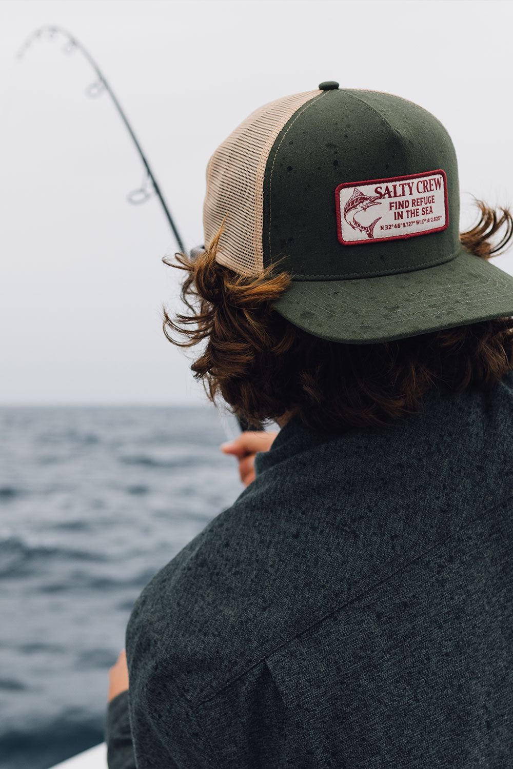Wearing the Salty Crew Coordinates Trucker in Deep Green, a person fishes from a boat, holding a rod over the ocean beneath an overcast sky.