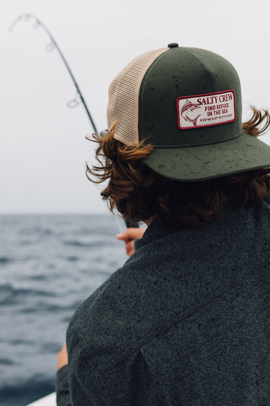 Wearing the Salty Crew Coordinates Trucker in Deep Green, a person fishes from a boat, holding a rod over the ocean beneath an overcast sky.