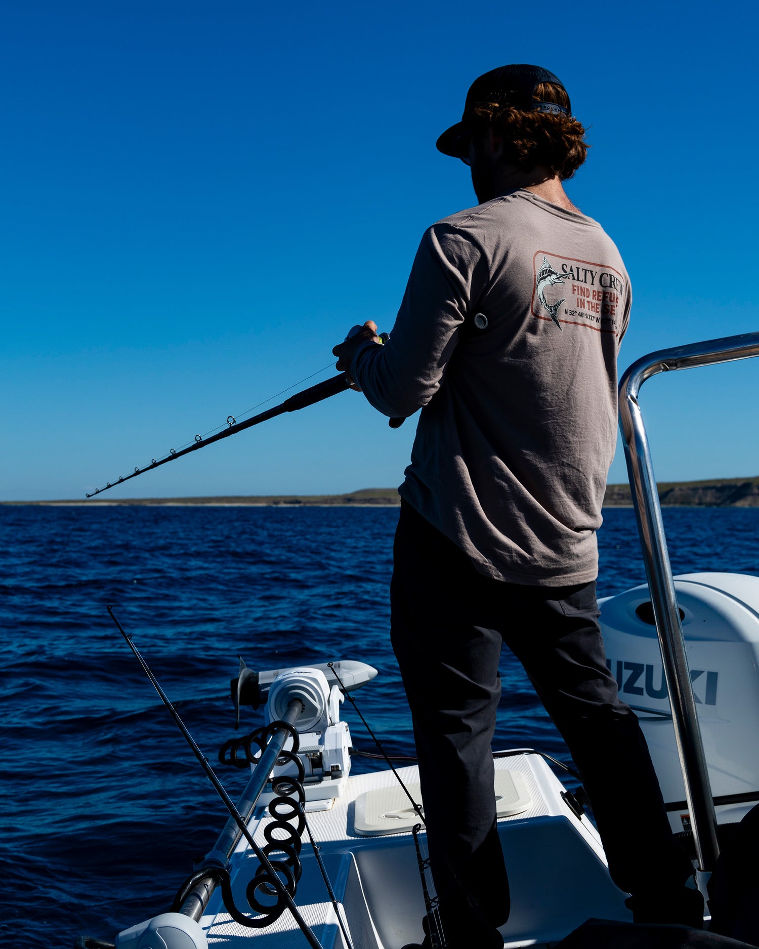 A person stands on a boat holding a fishing rod, facing the open blue ocean under a clear sky. Dressed in the Salty Crew Coordinates Crew Tee in dark grey, they are surrounded by fishing equipment and an outboard motor visible on the boat.