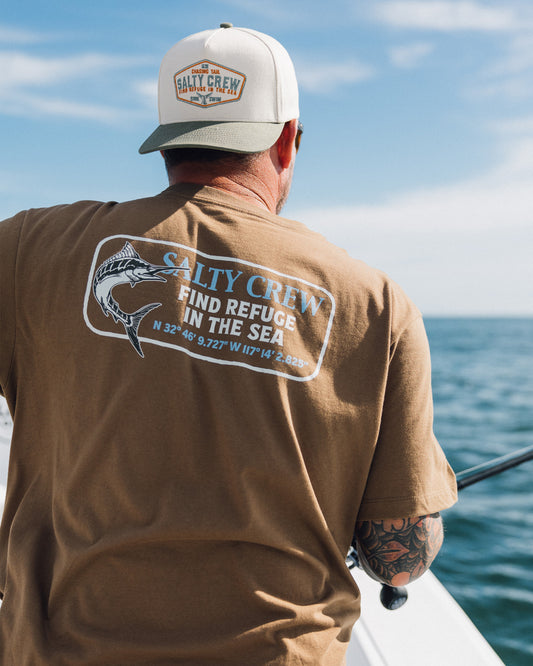 A person wearing a Salty Crew Coordinates Crew Tee in Sepia and a hat fishes on a boat. The cotton t-shirt features “Find refuge in the sea” text, coordinates, and a fish graphic. Ocean and sky can be seen in the background.