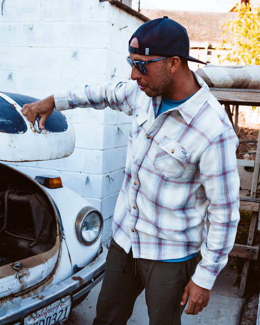 Wearing the Salty Crew Daybreak Flannel in Tradewinds, a man in a baseball cap lifts the hood of an old white car outdoors, standing near a white brick wall.