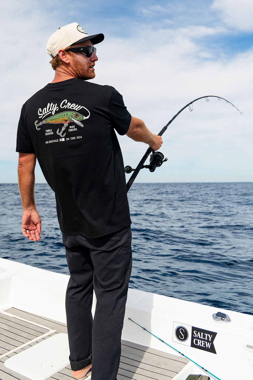 Wearing sunglasses and a black Salty Crew Decoy Crew Tee, a man fishes from a boat, holding his fishing rod over the open ocean beneath a clear sky.