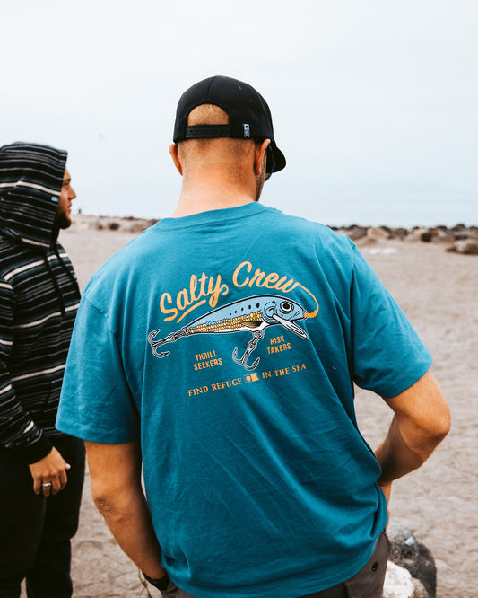A man stands on a rocky beach under an overcast sky, facing away, wearing the Salty Crew Decoy Crew Tee in Slate—crafted from soft, combed ringspun 100% cotton. Nearby, another person wears a striped hoodie.