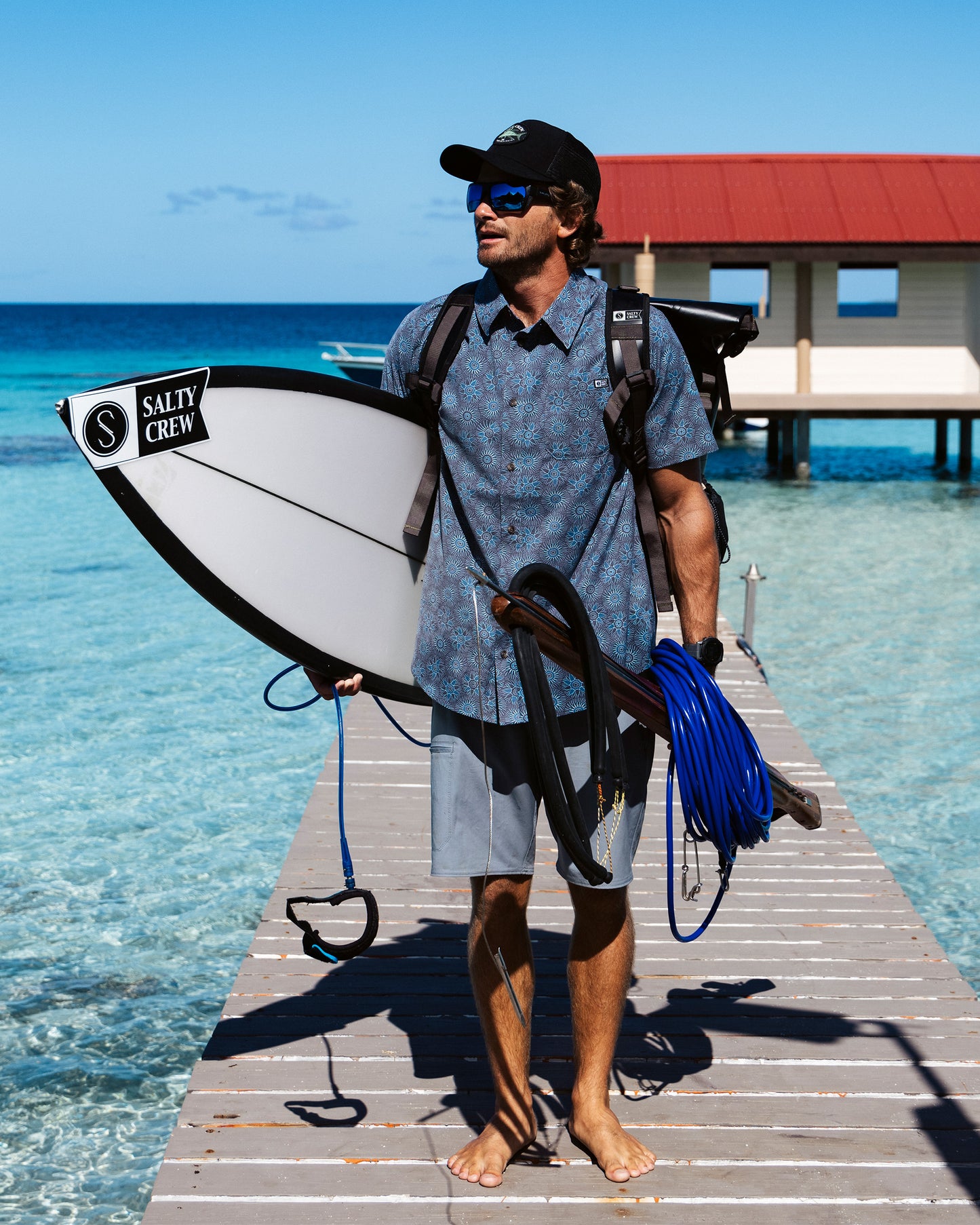 A man in the Salty Crew Drifter UV Button Up - Monument, sunglasses, and cap stands barefoot on a wooden dock with a surfboard and gear, clear blue water beneath him and a red-roofed building in the background.