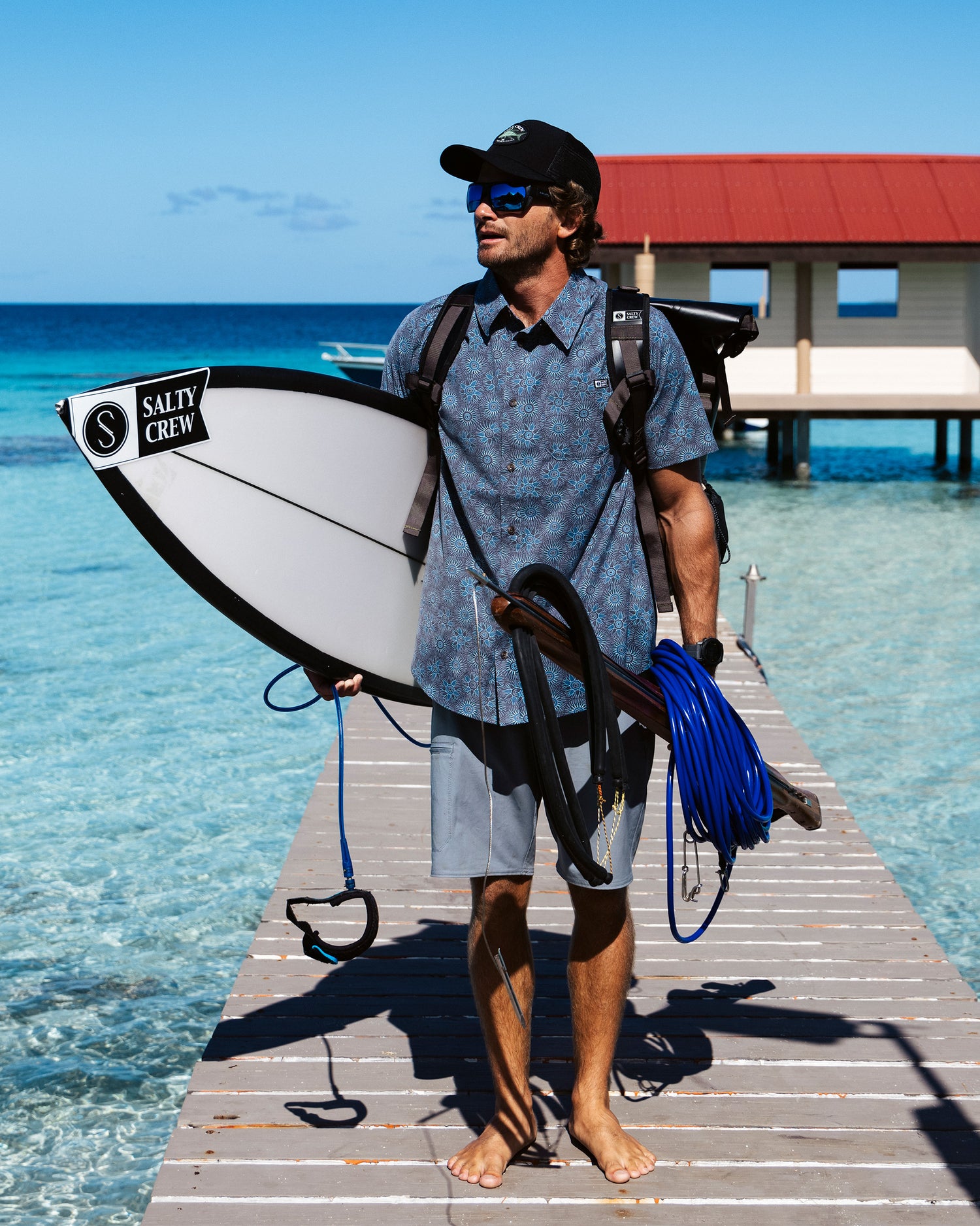 A man in the Salty Crew Drifter UV Button Up - Monument, sunglasses, and cap stands barefoot on a wooden dock with a surfboard and gear, clear blue water beneath him and a red-roofed building in the background.