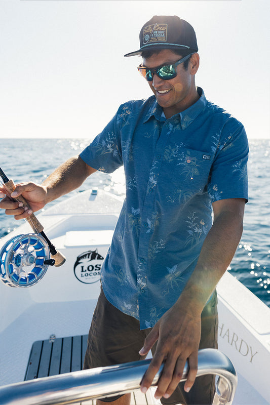 A man wearing the Salty Crew Drifter UV Button Up - Navy, featuring UPF 50+ sun protection, along with sunglasses and a cap, smiles while holding a fishing rod on a boat under the sunny sky. The ocean and bright sky create a vibrant backdrop.