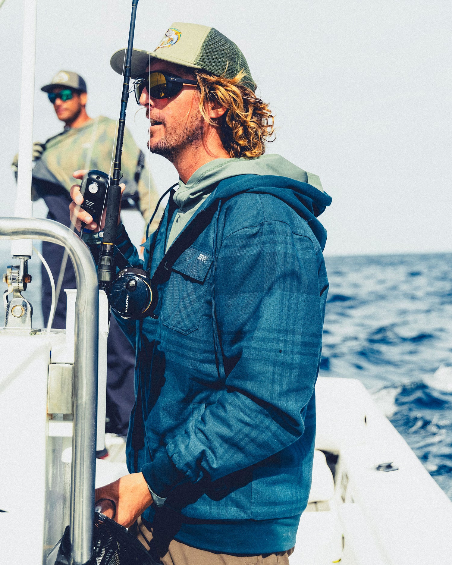 A man wearing a Salty Crew Fathom Jacket - Mid Blue and sunglasses holds a fishing rod on a boat at sea.