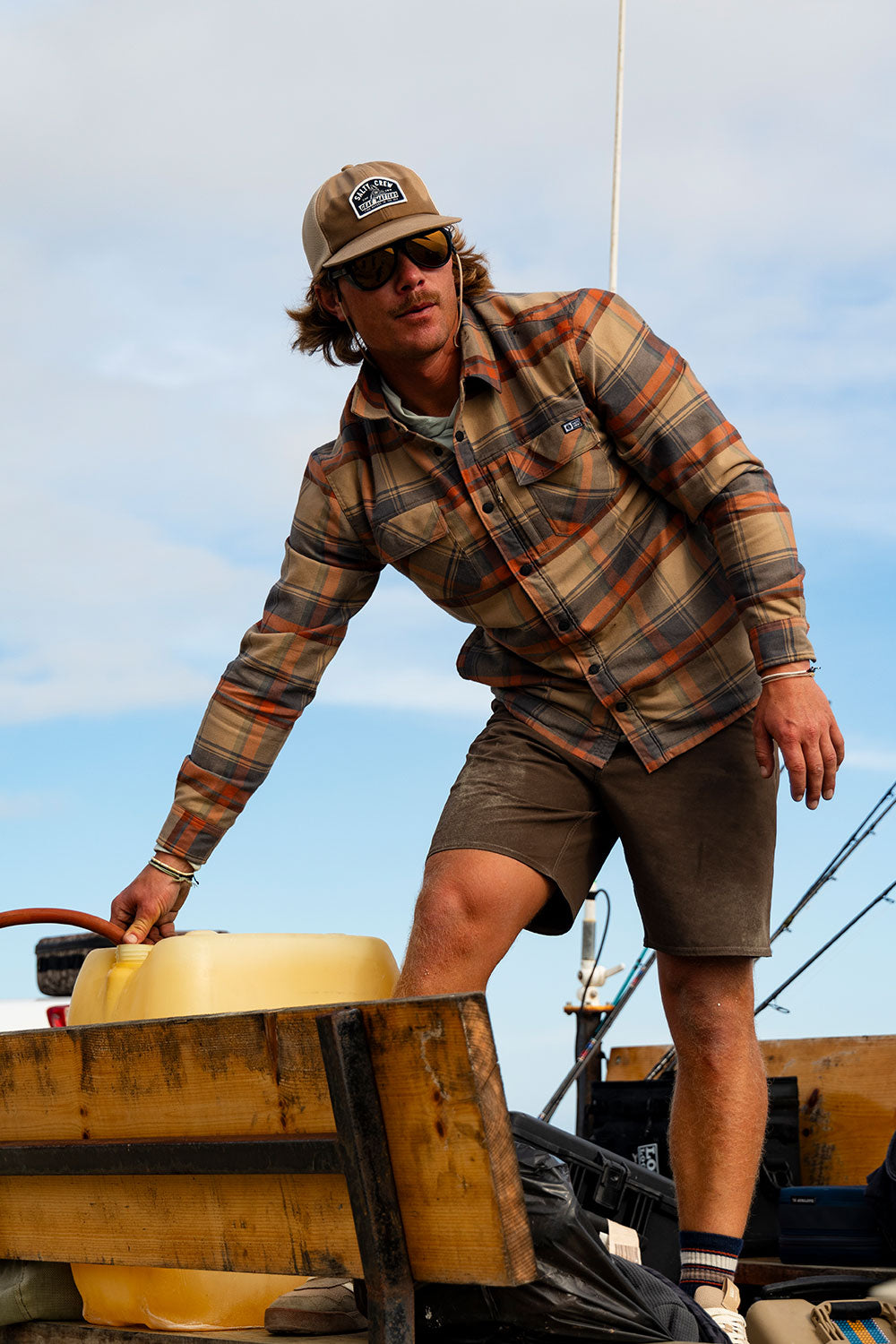 A man stands outdoors on a wooden surface, holding a yellow container, wearing the Salty Crew Fathom Tech Shacket in Twig along with mid-weight twill shorts featuring a cozy polar fleece liner and a cap. Fishing rods and blue sky can be seen in the background.