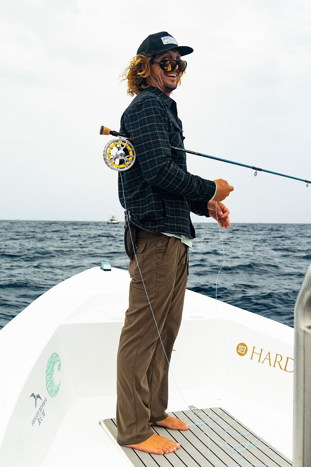 A person with long hair and sunglasses stands barefoot on a boat, wearing the Salty Crew Fathom Tech Flannel in Black Grey, holding a fishing rod over their shoulder, with the ocean and a distant boat in the background under a cloudy sky.