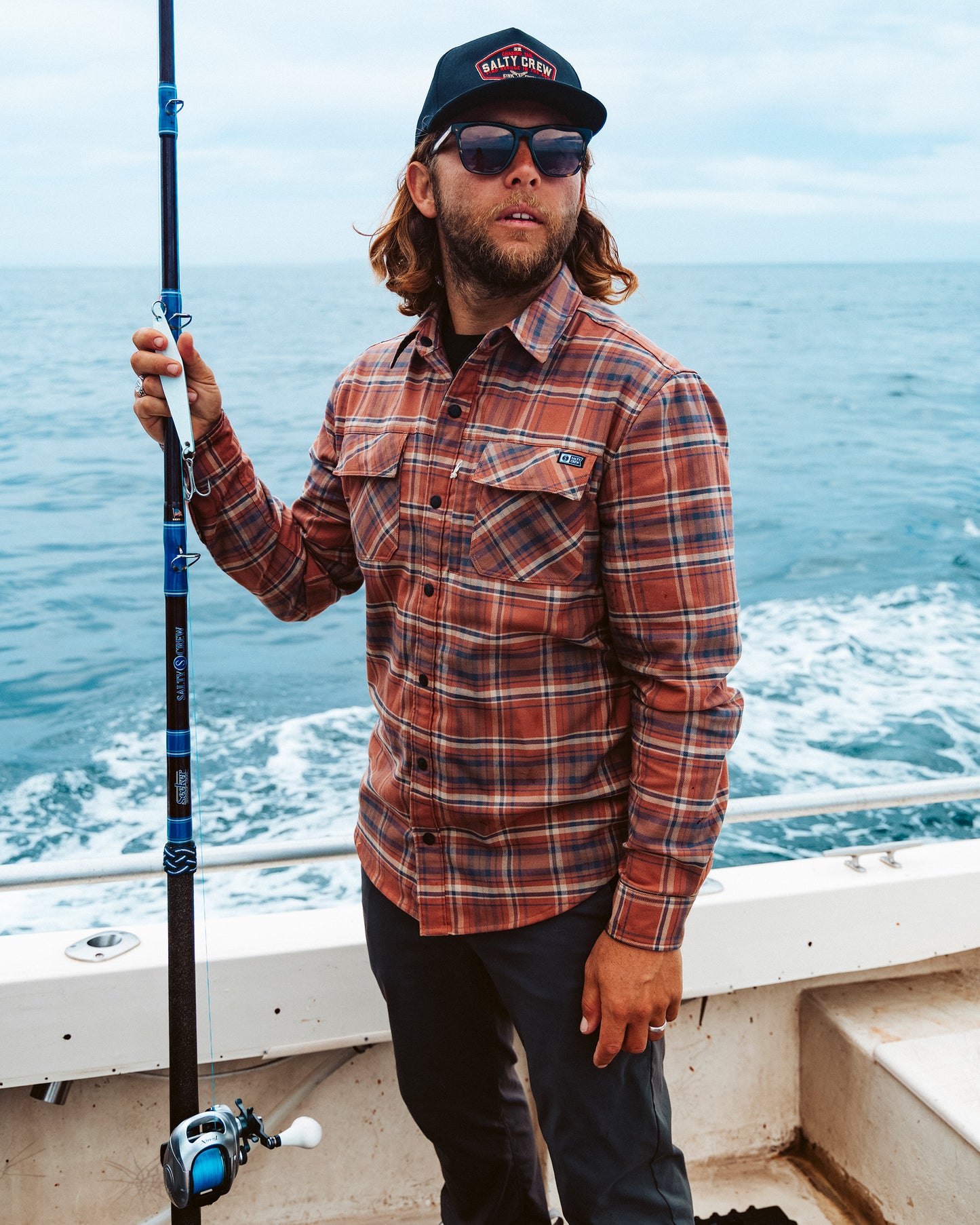 A man wearing the Salty Crew Fathom Tech Flannel in Russet and a hat holds a fishing rod on a boat, with the ocean in the background.
