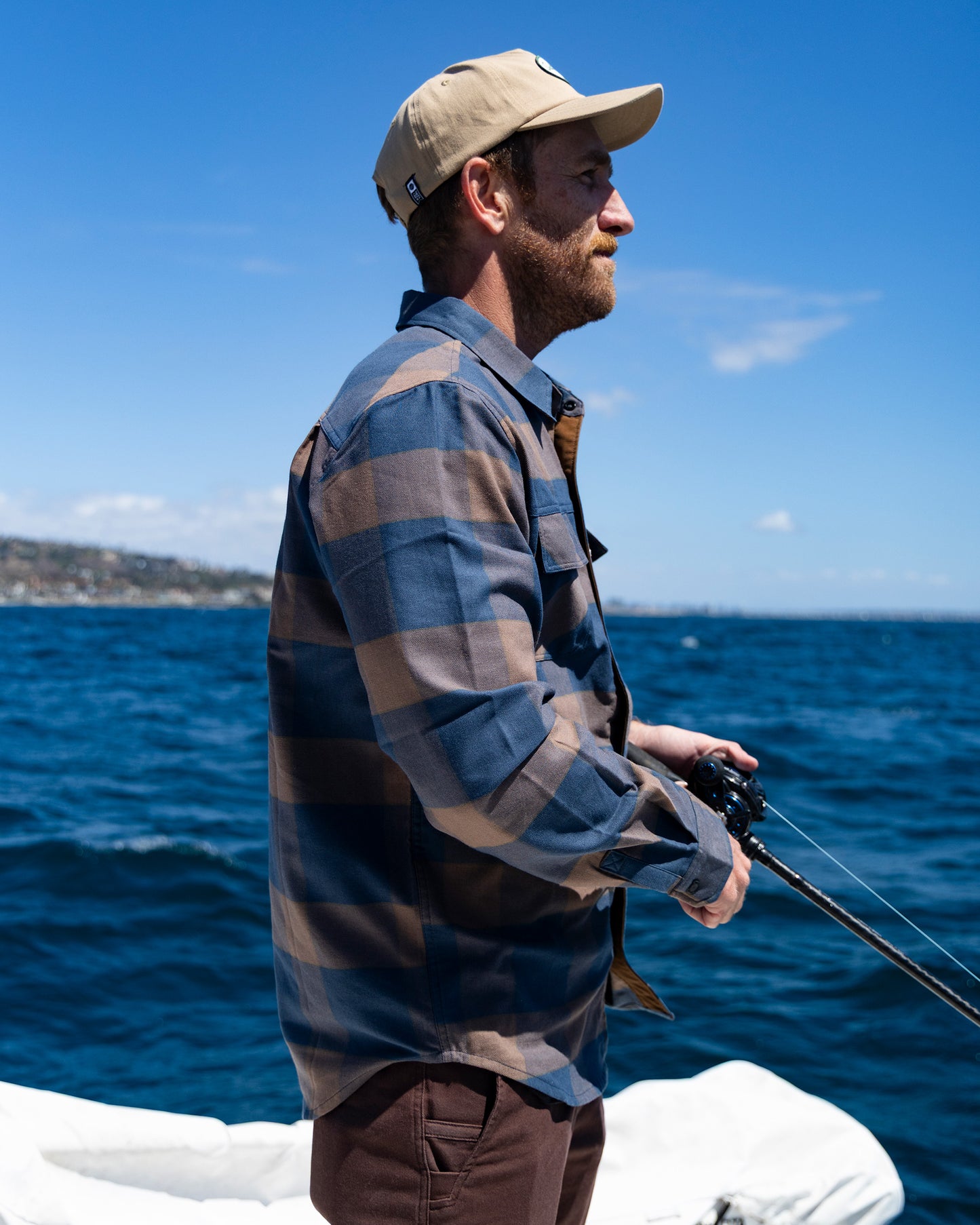 Wearing the Salty Crew Fathom Tech Flannel in Navy/Rust and a tan cap, a man fishes from a boat on blue ocean waters, with a distant shoreline visible beneath a clear, sunny sky.