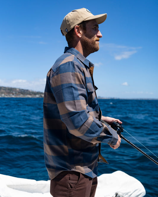 Wearing the Salty Crew Fathom Tech Flannel in Navy/Rust and a tan cap, a man fishes from a boat on blue ocean waters, with a distant shoreline visible beneath a clear, sunny sky.