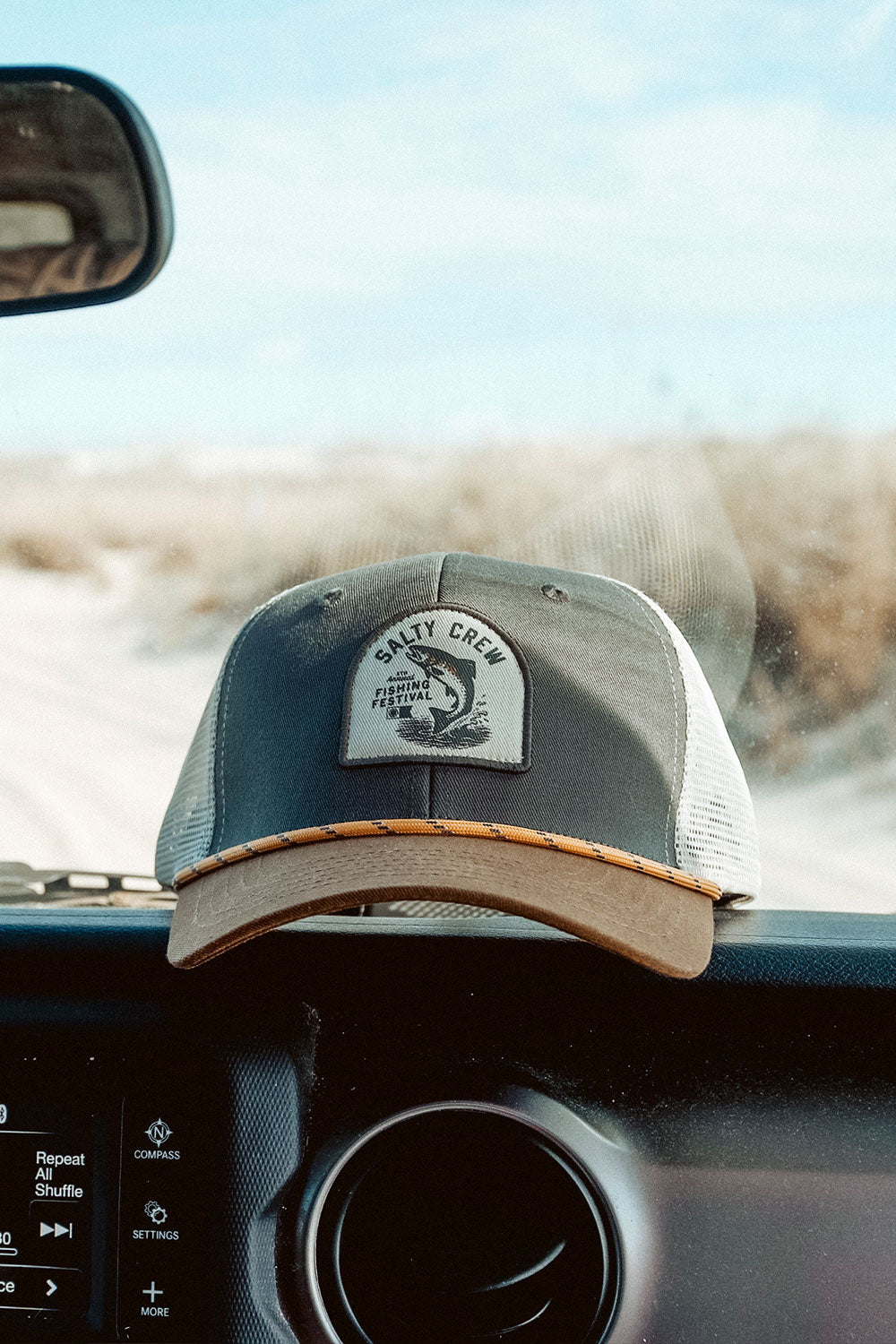 A Salty Crew Fishing Club Trucker hat in grey and brown, featuring a woven patch logo, rests on the dashboard of a car with sand dunes and blue sky visible through the windshield. This low-crown mesh trucker hat offers a comfortable cotton snapback for an adjustable fit.