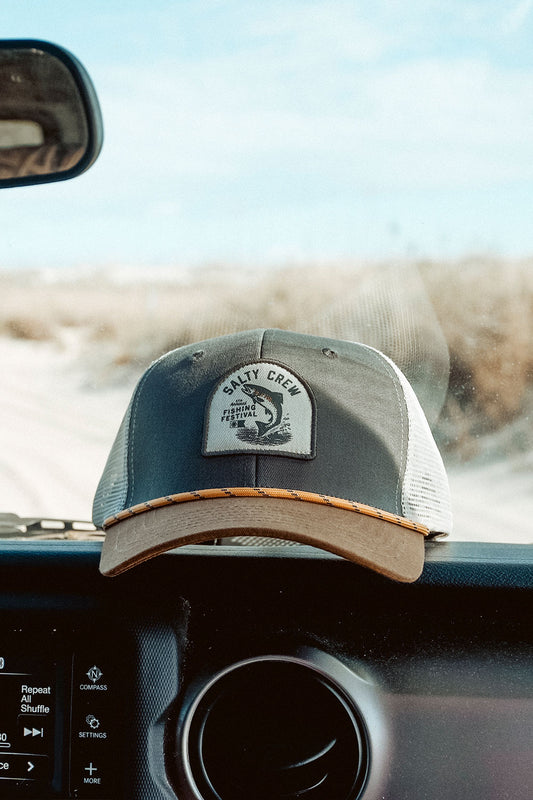 A Salty Crew Fishing Club Trucker hat in grey and brown, featuring a woven patch logo, rests on the dashboard of a car with sand dunes and blue sky visible through the windshield. This low-crown mesh trucker hat offers a comfortable cotton snapback for an adjustable fit.