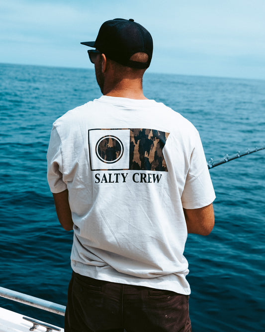 Wearing a Salty Crew Flagship Crew Tee in Bone, a person with a black cap stands on a boat fishing in the ocean under clear skies, their back to the camera with blue water and sky as the backdrop.
