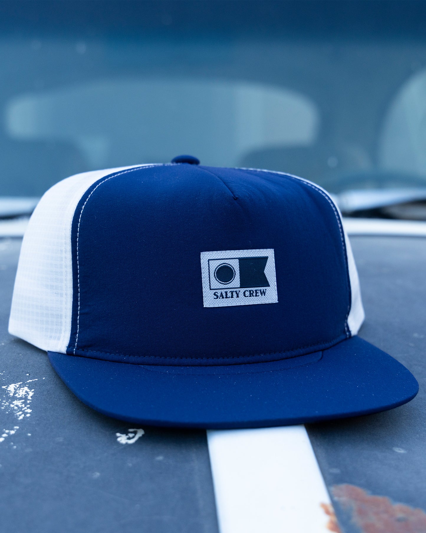 A Salty Crew Flagship Tech Trucker - Navy/Grey unstructured 5-panel hat with a branded patch rests on a weathered car hood marked by rust spots and a white stripe.