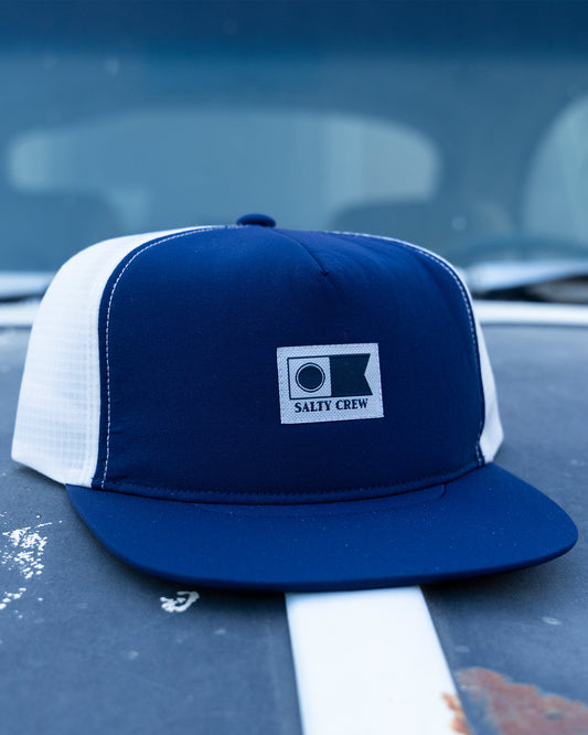 A Salty Crew Flagship Tech Trucker - Navy/Grey unstructured 5-panel hat with a branded patch rests on a weathered car hood marked by rust spots and a white stripe.