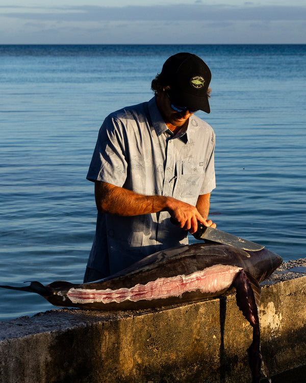 Wearing the Salty Crew Flagship UV Button Up - Tradewinds, a person fillets a large fish on a concrete ledge by the calm ocean under a clear sky, sunlight casting shadows as they work.