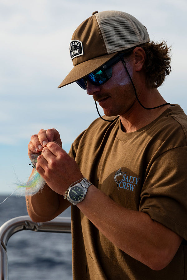 Man on a boat wearing a khaki Gear Shop Trucker hat and Salty Crew shirt while tying a fishing lure