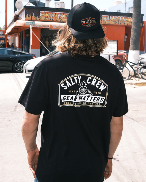 Wearing a black Salty Crew Gear Shop Original Tee—crafted from 100% cotton and screenprinted for lasting quality—a person with long hair stands with their back to the camera on a sunny street near an orange restaurant.