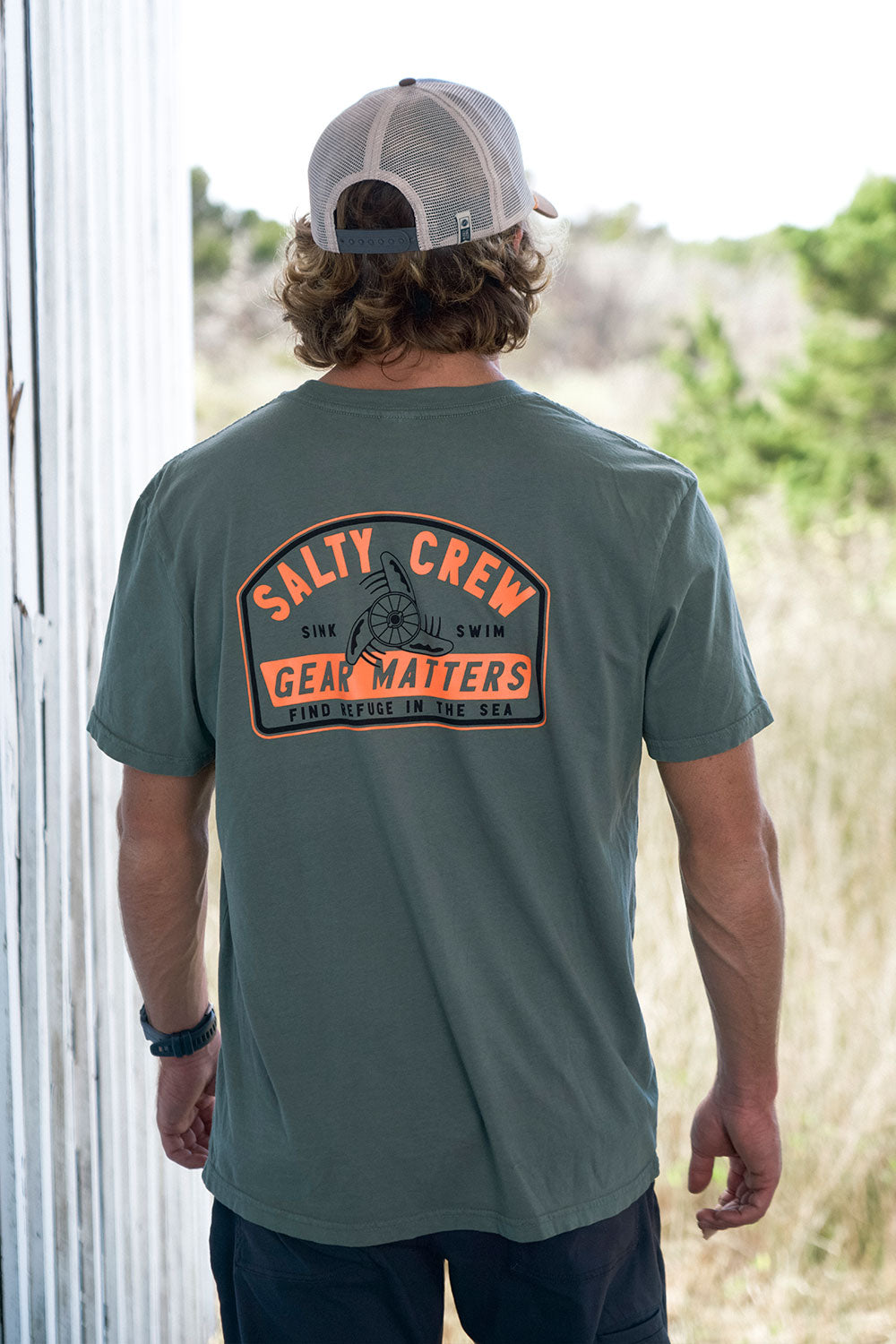 A person wearing the Salty Crew Gear Shop Original Tee in Laurel Wreath and a matching cap stands outdoors near a white wall, with greenery in the background. The soft spun cotton shirt features a screenprinted fishing graphic and the words “Sink Swim Gear Matters Find Refuge In The Sea.”.