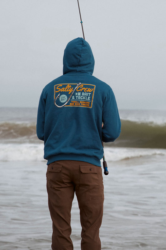 A person wearing the Salty Crew Hooked Up Fleece in Cast Blue, a mid-weight cotton polyester hoodie with a kangaroo pocket, stands on the shore fishing with a rod. They pair the hoodie with brown pants as waves roll in behind them.