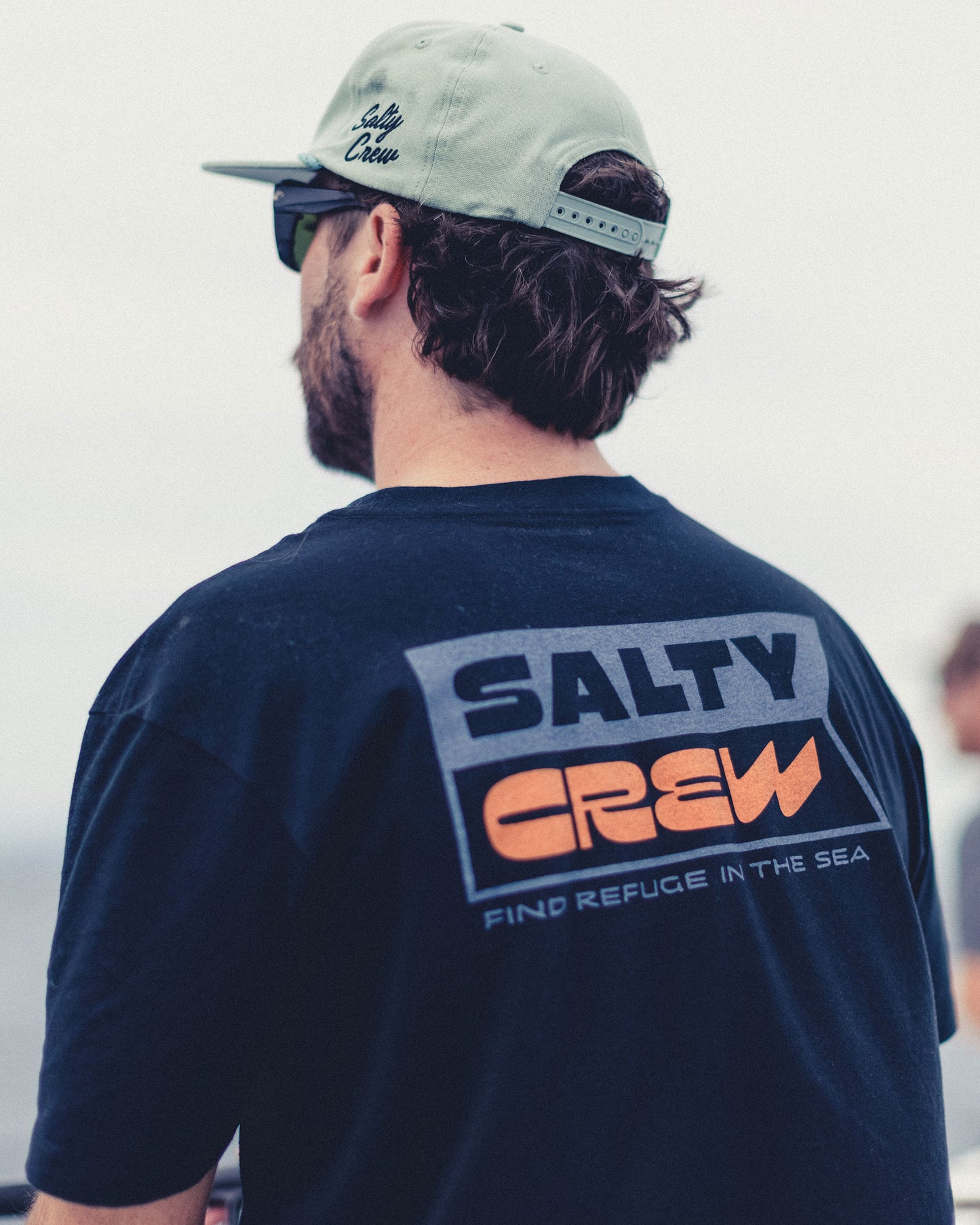 A man wearing a black Inverse Flag Crew Tee, a 100% cotton short-sleeved shirt with a screenprinted rectangular graphic on the back that reads SALTY CREW in large text and FIND REFUGE IN THE SEA below it.
